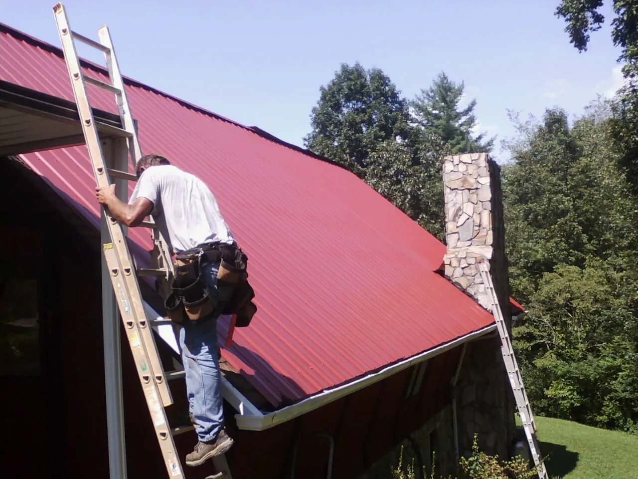 Roofer on a ladder beside a red metal roof and stone chimney — Soffit & Fascia Repair by Roofing Brothers in Pasadena